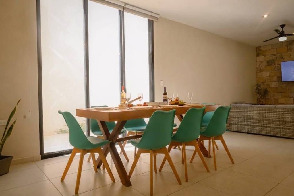 A dining area with a wooden table and four chairs, a television mounted on a stone wall, and a large window with blinds.
