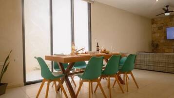 A dining area with a wooden table and four chairs, a television mounted on a stone wall, and a large window with blinds.