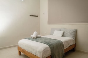 Ein ordentlich bezogenes Bett mit weißen Bettwäsche und einer grünen Tagesdecke, ein hölzernes Kopfende und eine wandmontierte Leuchte.
