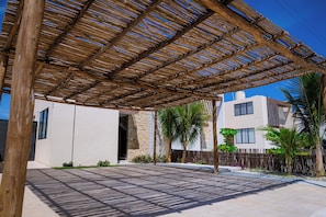 Ein überdachter Außenbereich mit einer Holz-Pergola, einer gepflasterten Terrasse und Blick auf ein modernes Gebäude mit Steinaußenwand.