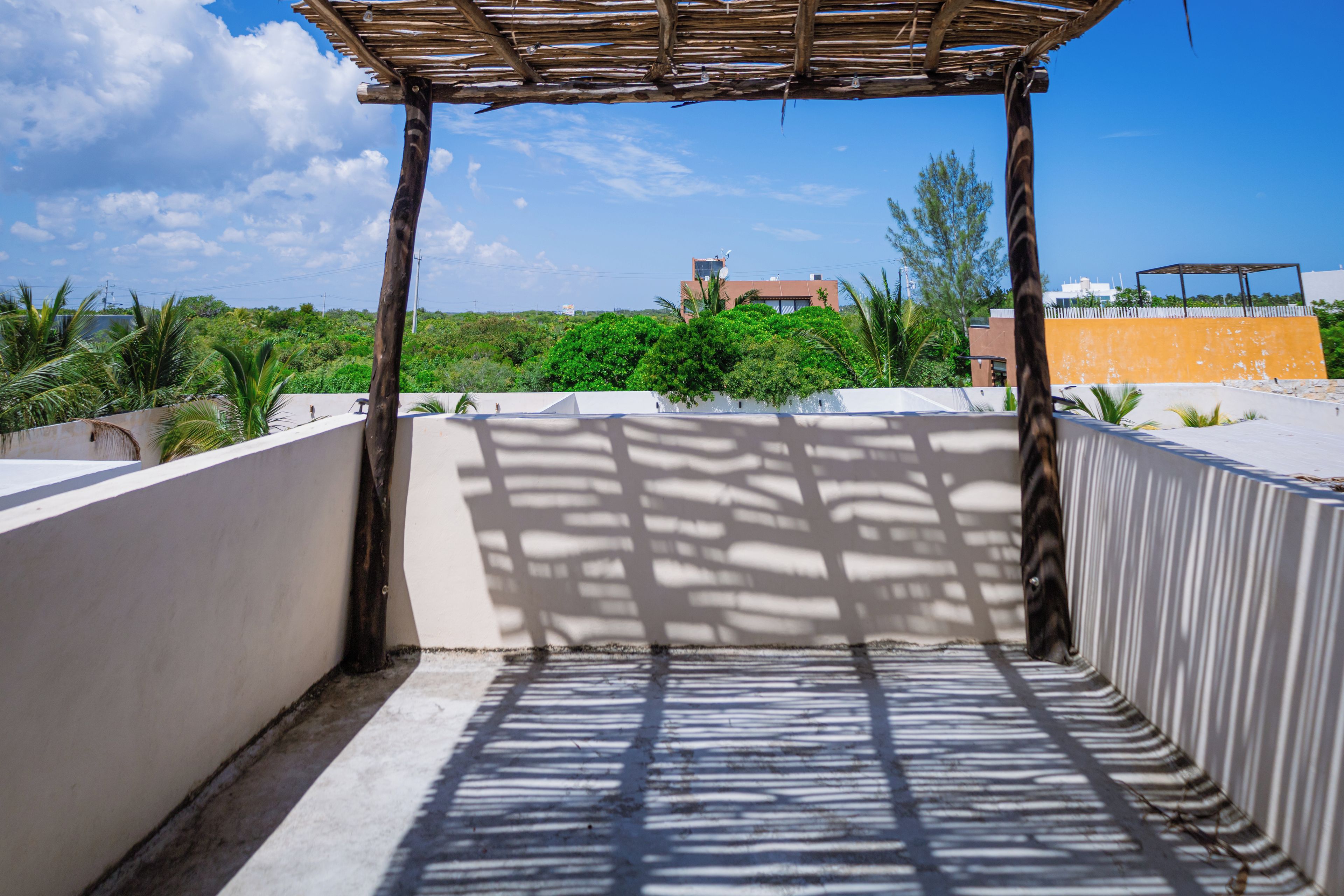 A rooftop terrace with a pergola structure, a view of greenery and buildings in the background.