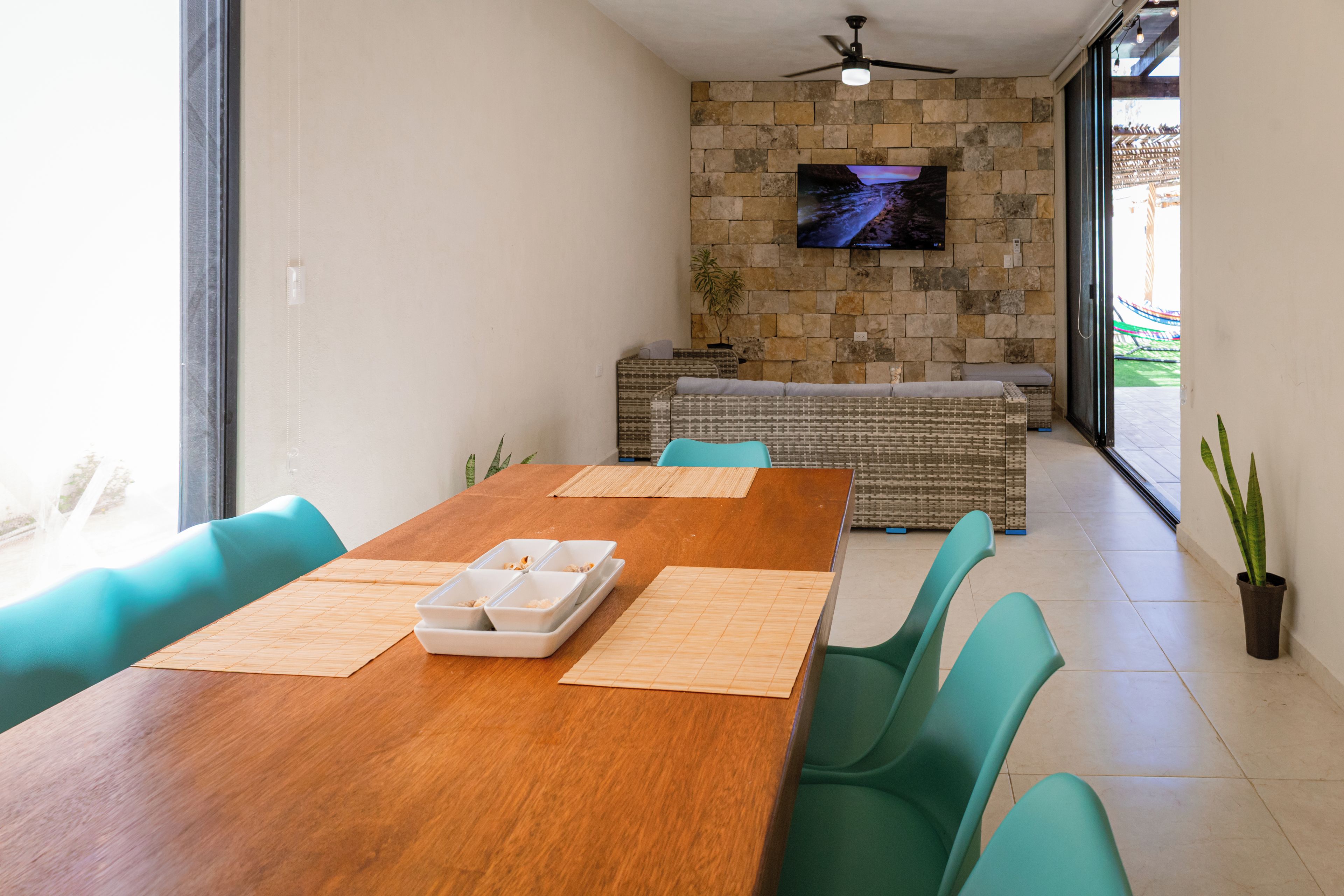 A dining area with a wooden table, green chairs, a stone wall with a TV, and a view through glass doors.