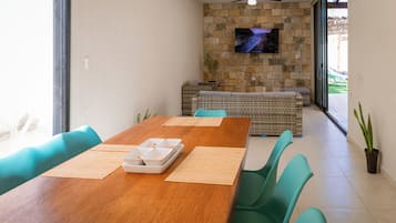 A dining area with a wooden table, green chairs, a stone wall with a TV, and a view through glass doors.
