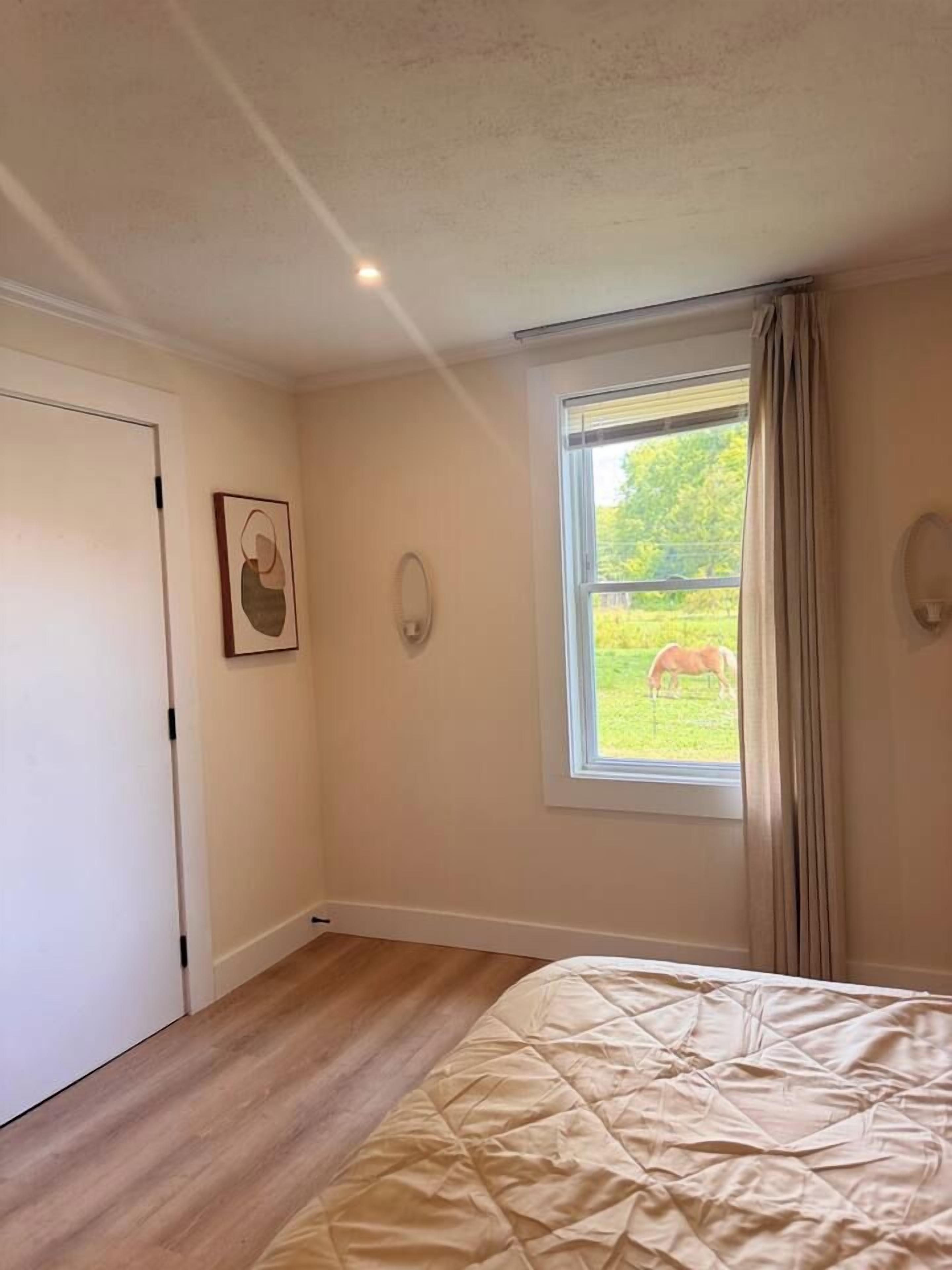 Un dormitorio con cama, ventana con vista a un caballo y puerta.