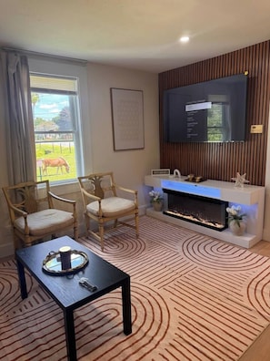 Una sala de estar con chimenea, dos sillas de madera, una mesa de centro y un televisor de pantalla plana montado en la pared.