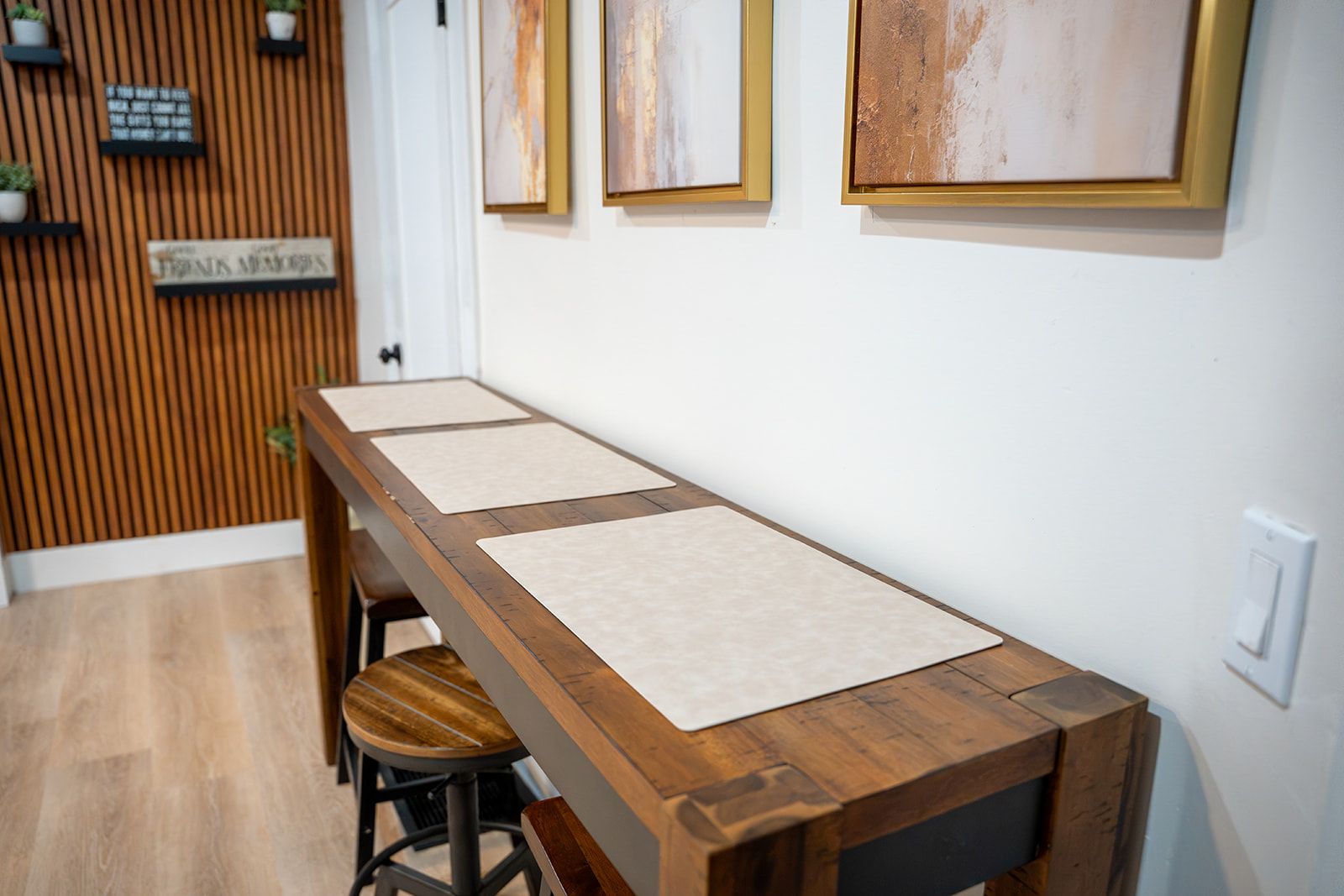 A long wooden dining table with three beige placemats, a wooden slatted wall, and a single bar stool.