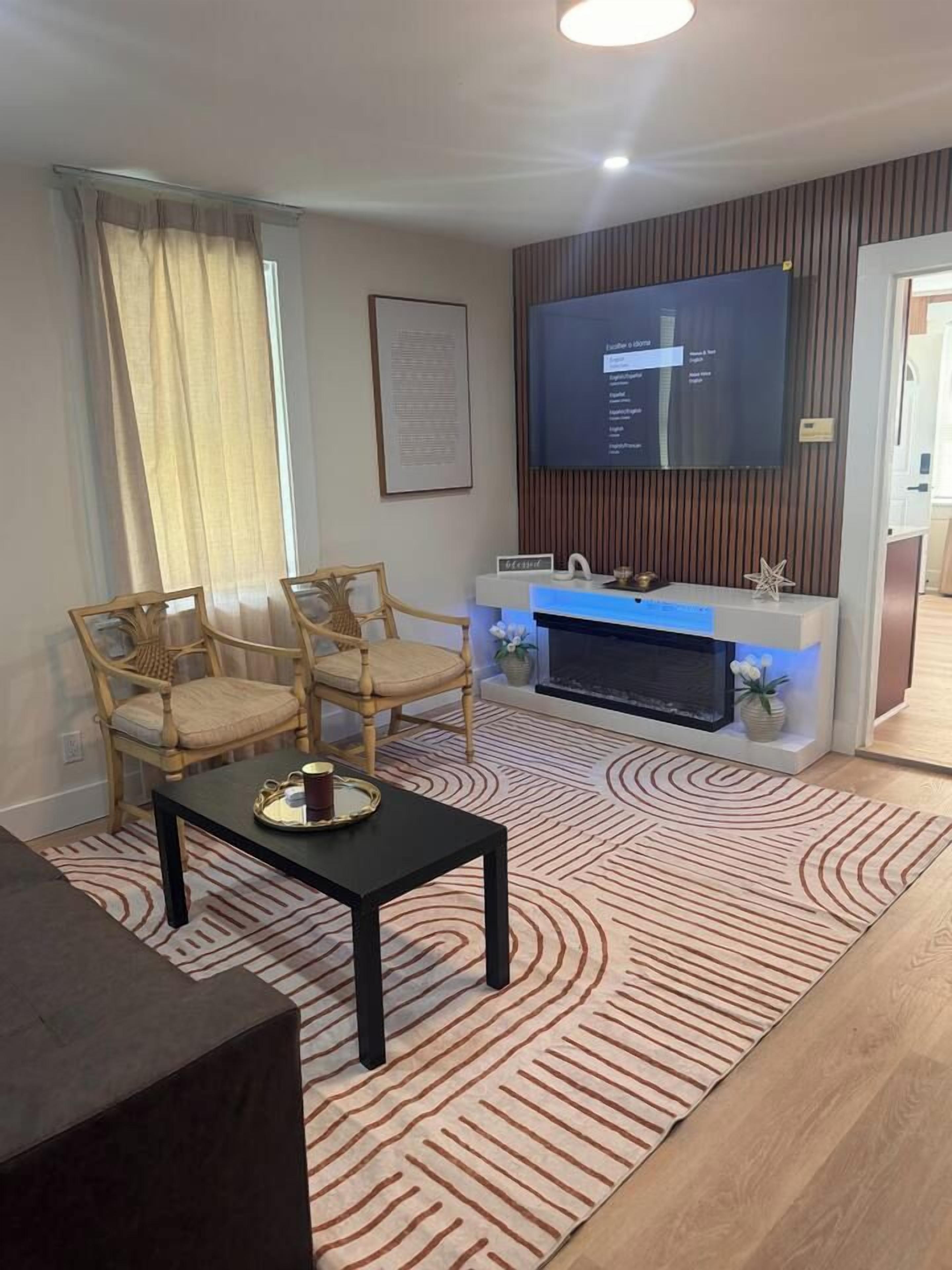 Un salon équipé d’un téléviseur à écran plat, de deux fauteuils en bois, d’une table basse noire et d’un tapis à motifs.
