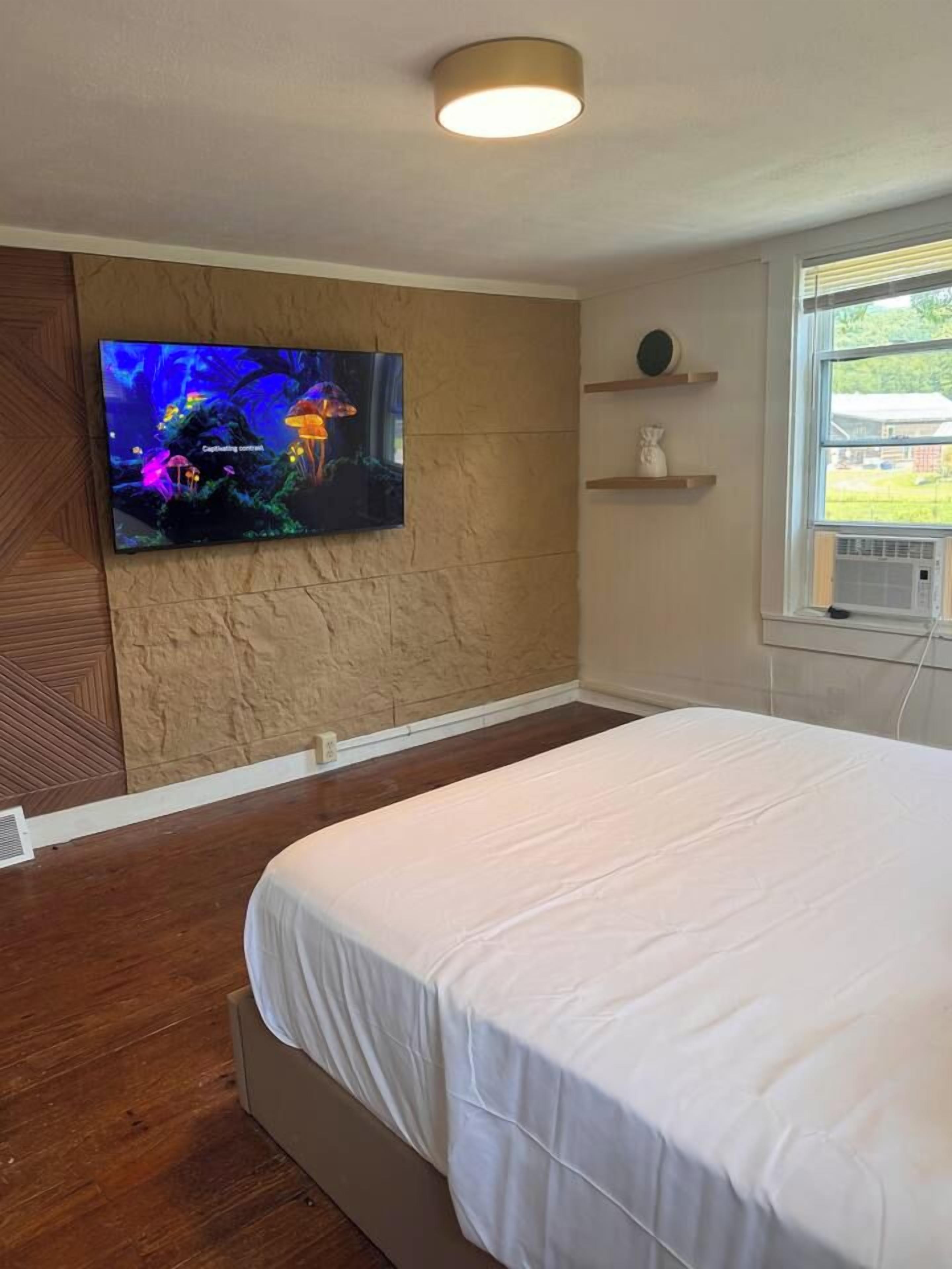 Een slaapkamer met een bed, een aan de muur bevestigde televisie die een onderwaterscène toont, een raam met uitzicht op groen en een wandplank met een vaas.