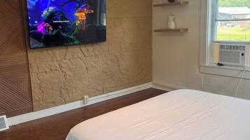 Een slaapkamer met een bed, een aan de muur bevestigde televisie die een onderwaterscène toont, een raam met uitzicht op groen en een wandplank met een vaas.
