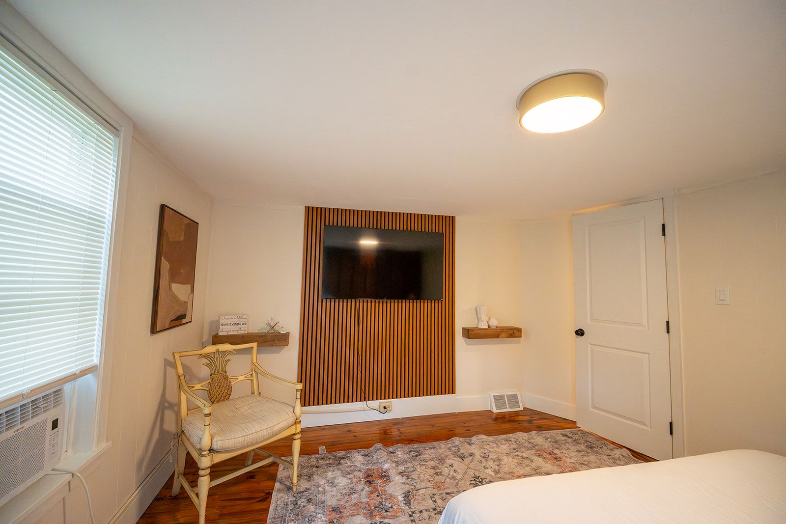 Un dormitorio con cama, una silla, un televisor montado en una pared de paneles de madera, un radiador y una puerta.