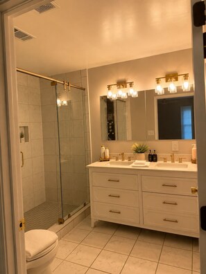 A modern bathroom with a glass shower enclosure, a vanity with a large mirror, and a sink with gold fixtures. -     30+ Day Stay - Travel Nurses & Work Crews (Tuscaloosa)