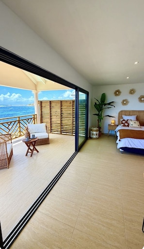 Ein Zimmer mit Balkon und Meerblick, ein Bett mit tropisch bedruckter Bettwäsche und ein Blumentopf.