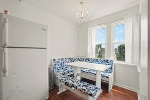 Eine kleine Küche mit einem weißen Kühlschrank, einer Bank mit blauem Blumenmuster, einem weißen Tisch und einem Fenster mit weißen Vorhängen.