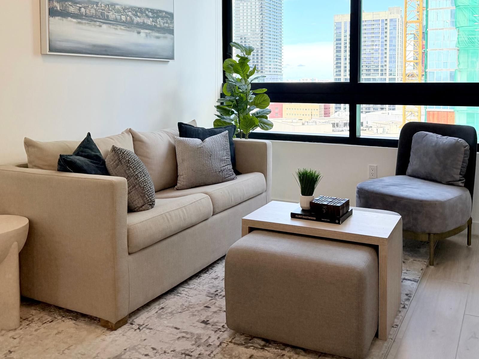 Una sala moderna con un sofá, un sillón y un reposapiés, una mesa de centro con un libro, una planta y vistas a la ciudad a través de amplios ventanales.