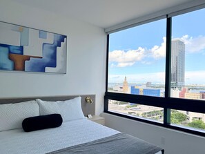 A hotel room with a large window, a bed with white bedding, a nightstand, and a painting on the wall. - Downtown Miami Luxe 1BR! (Miami)