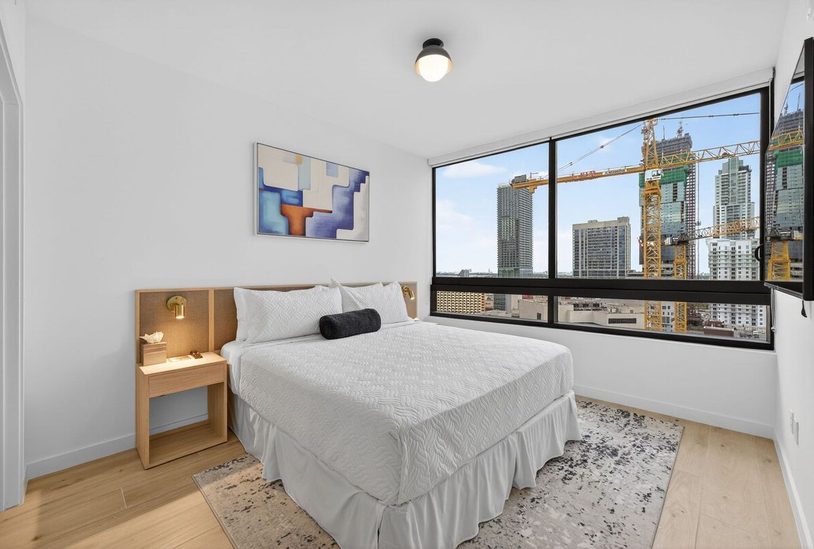 A bedroom with a large window, a bed, a nightstand, and a painting on the wall.
