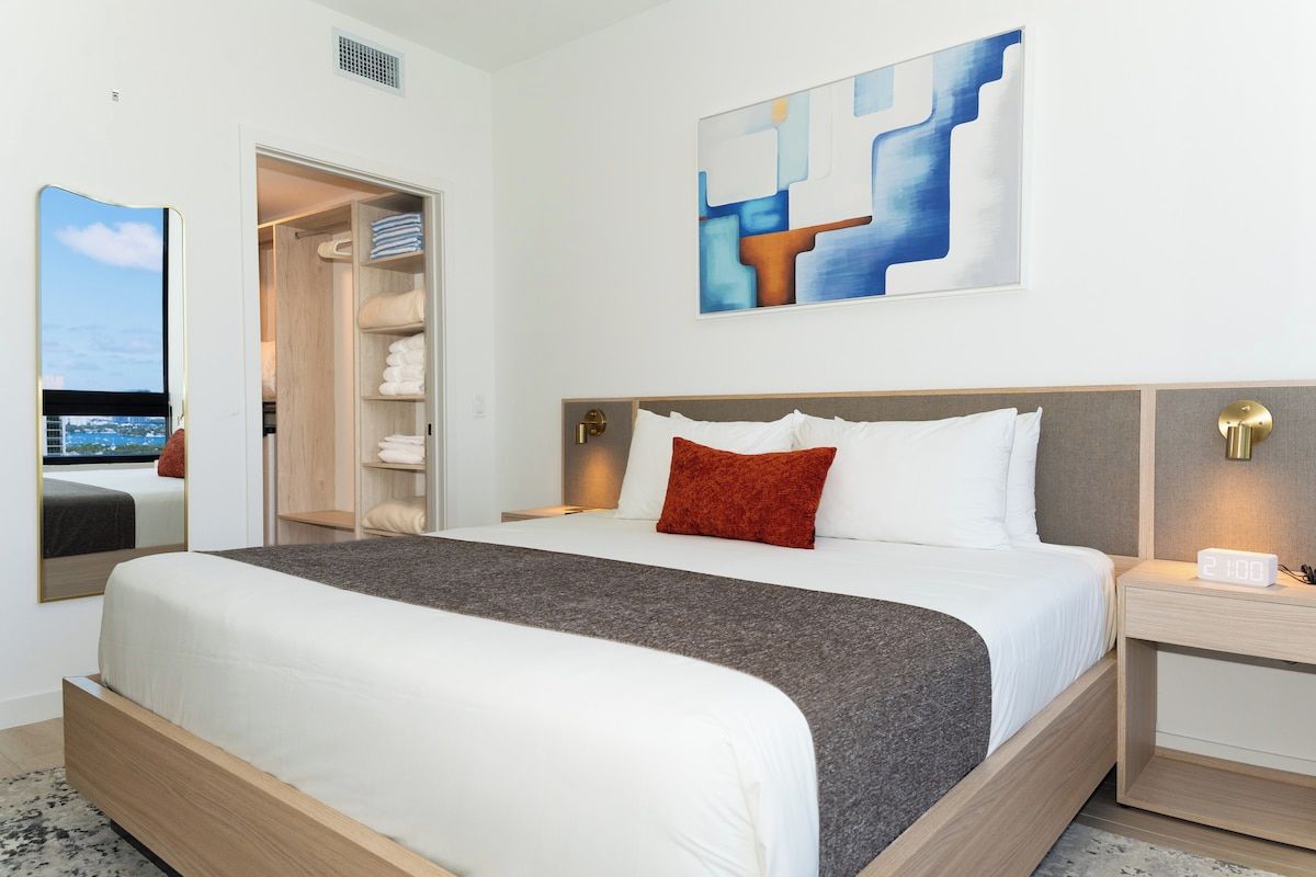 Quarto de hotel com uma cama grande, um guarda-roupa, uma mesa de cabeceira com relógio e um quadro na parede.