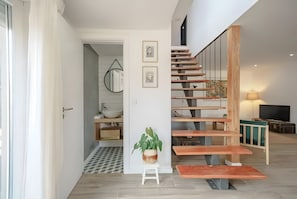 Ein modernes Interieur mit einer Holztreppe, einem kleinen weißen Hocker mit Pflanze und einem im Hintergrund sichtbaren Küchenbereich.