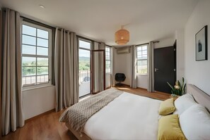 Ein Schlafzimmer mit einem großen Bett, einem Sessel, einem Schreibtisch und Blick ins Freie durch die Fenster.