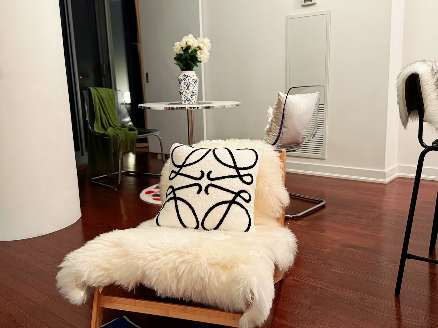 A modern living room with a white furry chair, a small round table with a vase of flowers, and a chair with a fluffy pillow.
