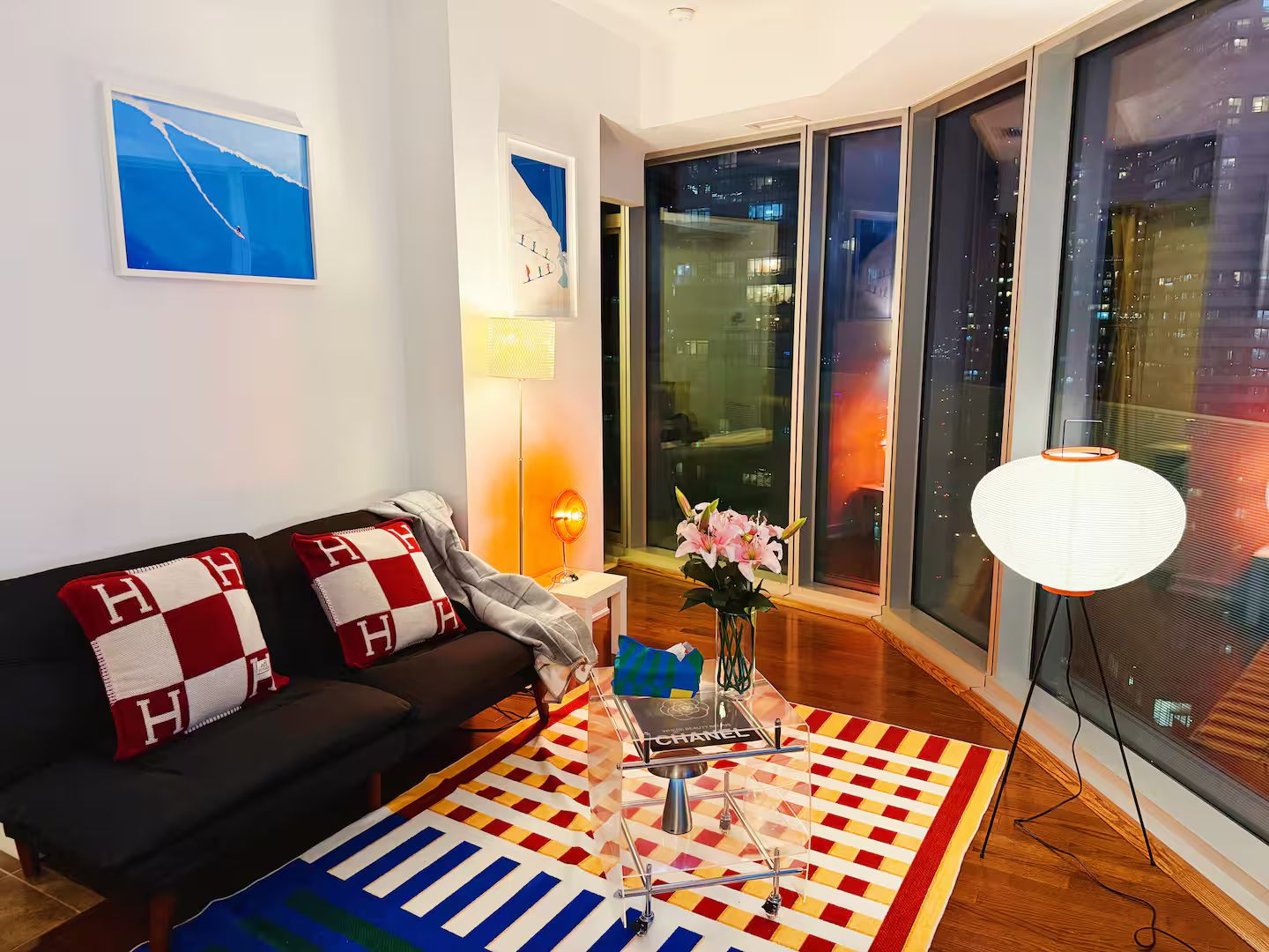 Un salon moderne avec un canapé, une lampe décorative, un vase de fleurs et une vue sur la ville grâce à de grandes fenêtres.