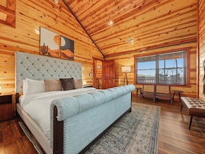 A bedroom with a large bed, a wooden ceiling, a window with blinds, and a small table with chairs. - Smoky Mtn Views | Indoor Pool, Hot Tub & Sauna (Sevierville)
