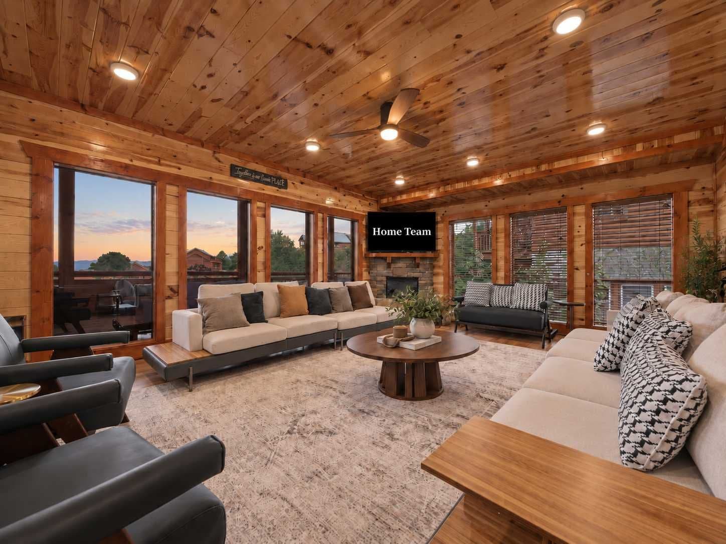 A spacious living room with a large sofa, a coffee table, and a fireplace. There is a television mounted on the wall and large windows offering a view of the outdoors.