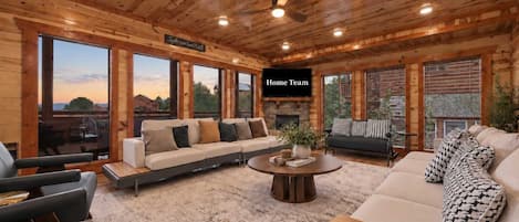 A spacious living room with a large sofa, a coffee table, and a fireplace. There is a television mounted on the wall and large windows offering a view of the outdoors.