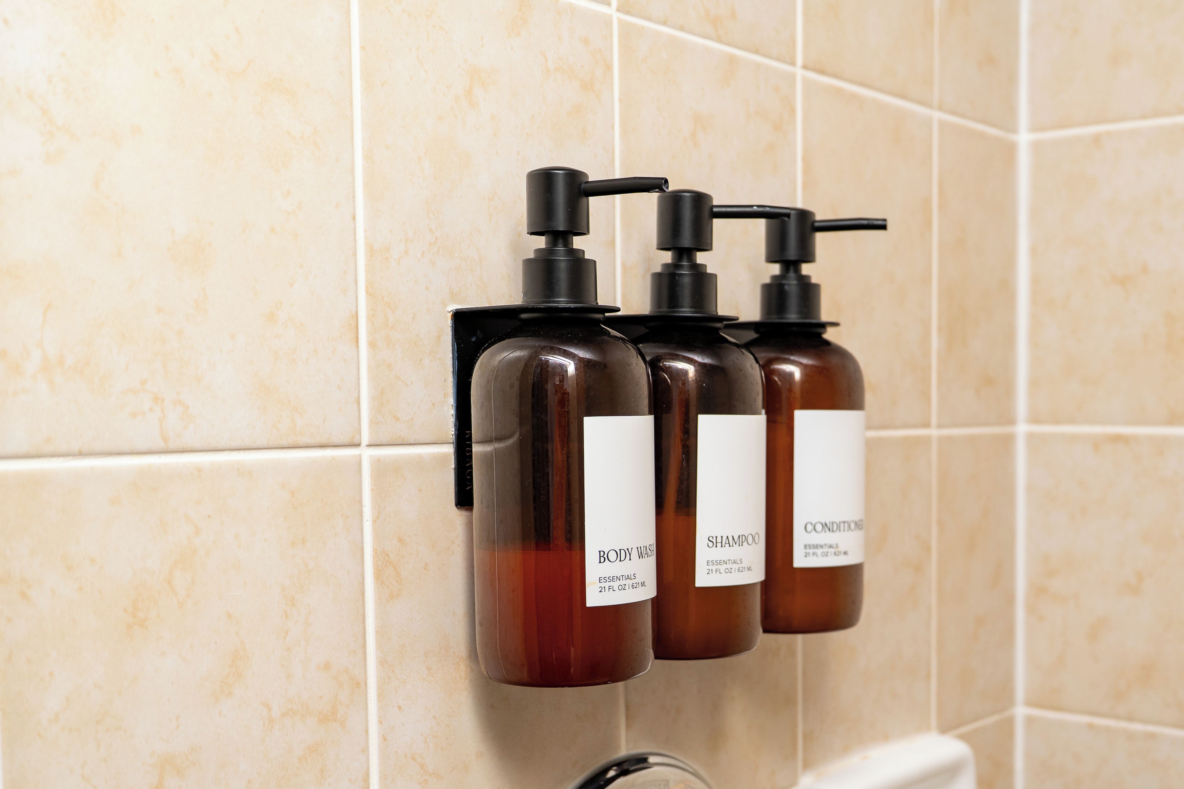 Three bottles of body wash and shampoo on a wall-mounted dispenser in a bathroom.