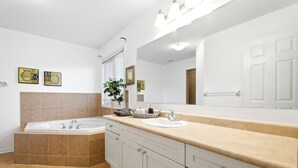 Un baño moderno con bañera, lavamanos y un espejo grande. Hay dos cuadros enmarcados en la pared y una planta en maceta sobre el mostrador.