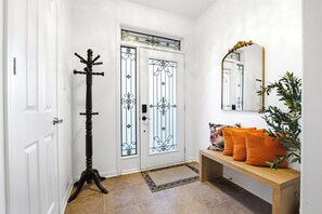 Un pasillo con una puerta blanca, un perchero, un banco con cojines y un espejo en la pared.