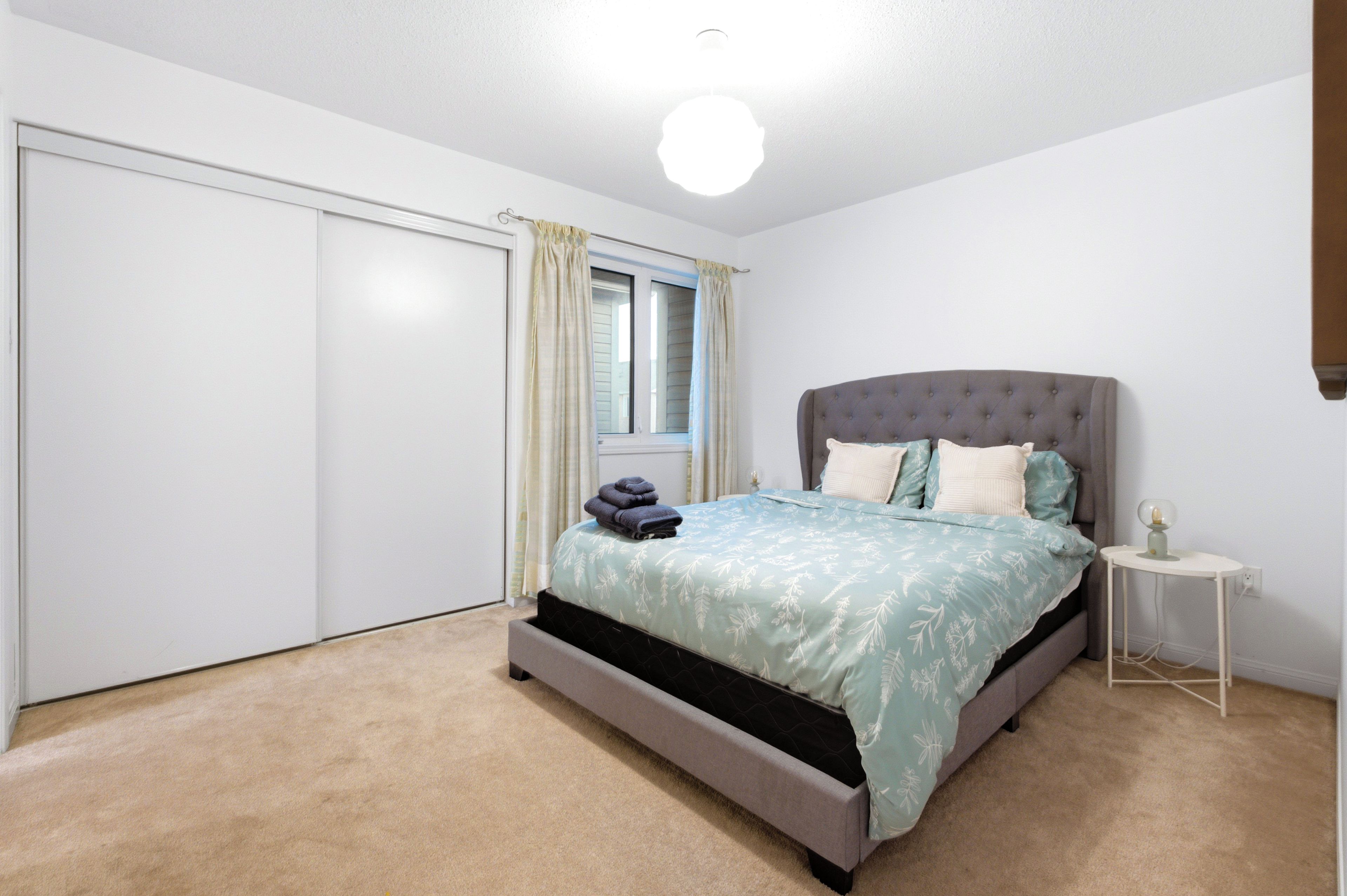 Une chambre à coucher avec un lit, une table de chevet, une fenêtre avec des rideaux et un luminaire au plafond.