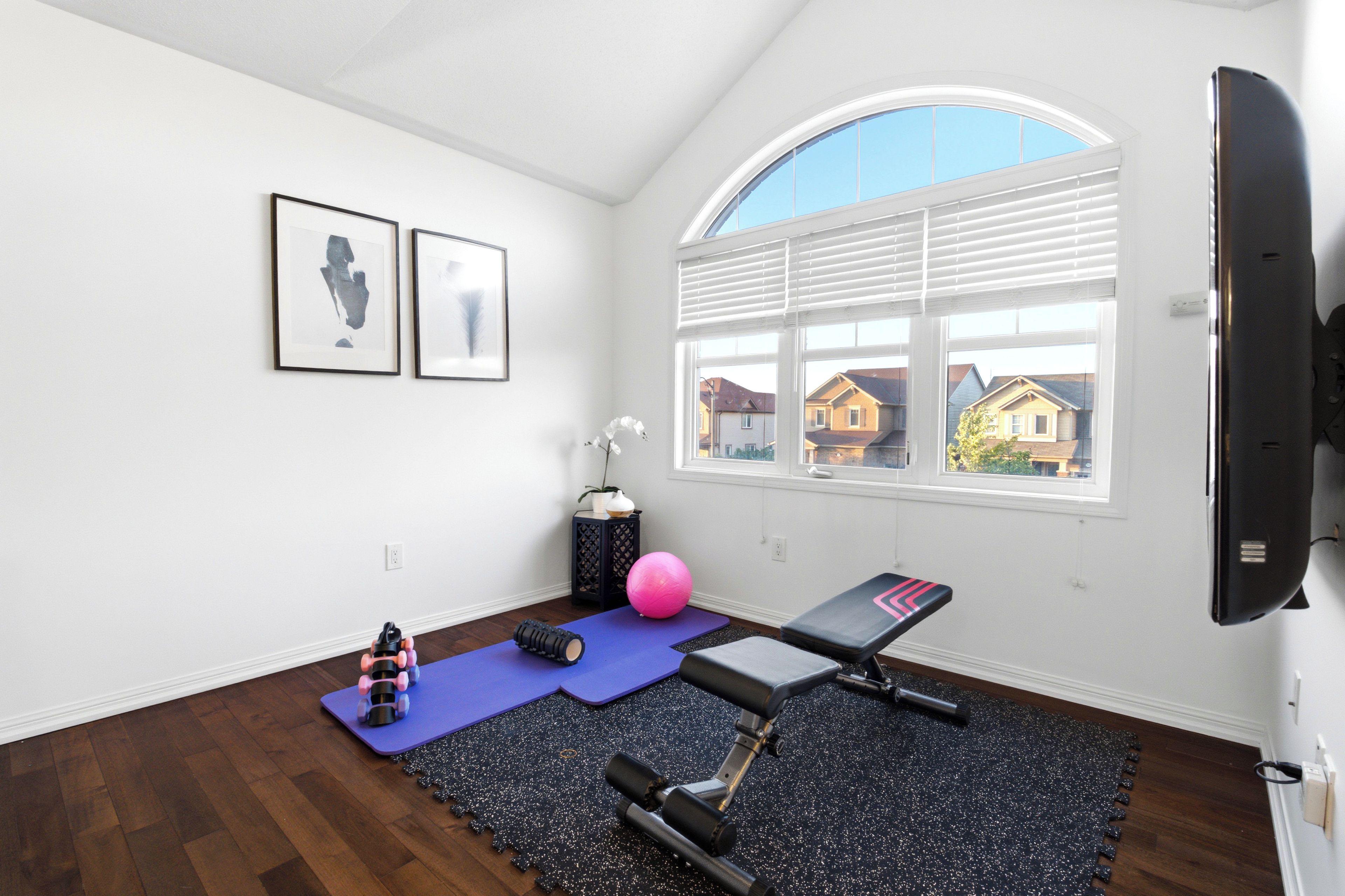 A room with a large window, a treadmill, a yoga mat, and a wall-mounted TV.