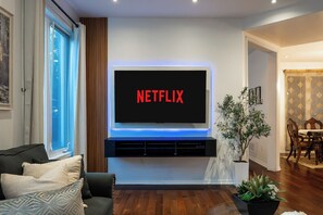 Una sala de estar con un televisor de pantalla plana montado en la pared, un sofá de color oscuro, una planta en maceta y un comedor al fondo.