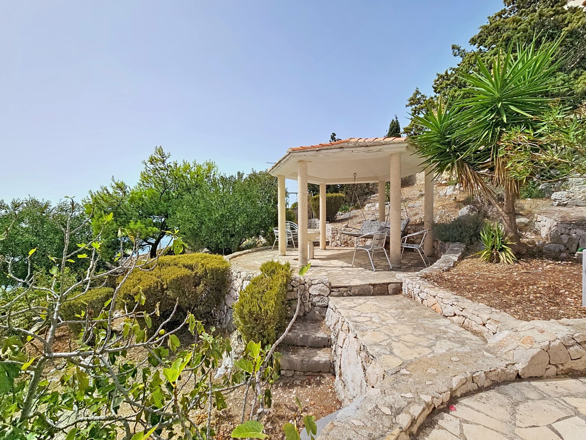 A stone pathway leading to a white gazebo with a tiled roof, surrounded by greenery and a view of the sea.