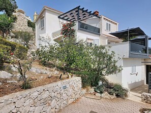 Ein modernes, zweistöckiges Haus mit einer Steinmauer, einer überdachten Außenfläche und einem Garten mit verschiedenen Pflanzen.