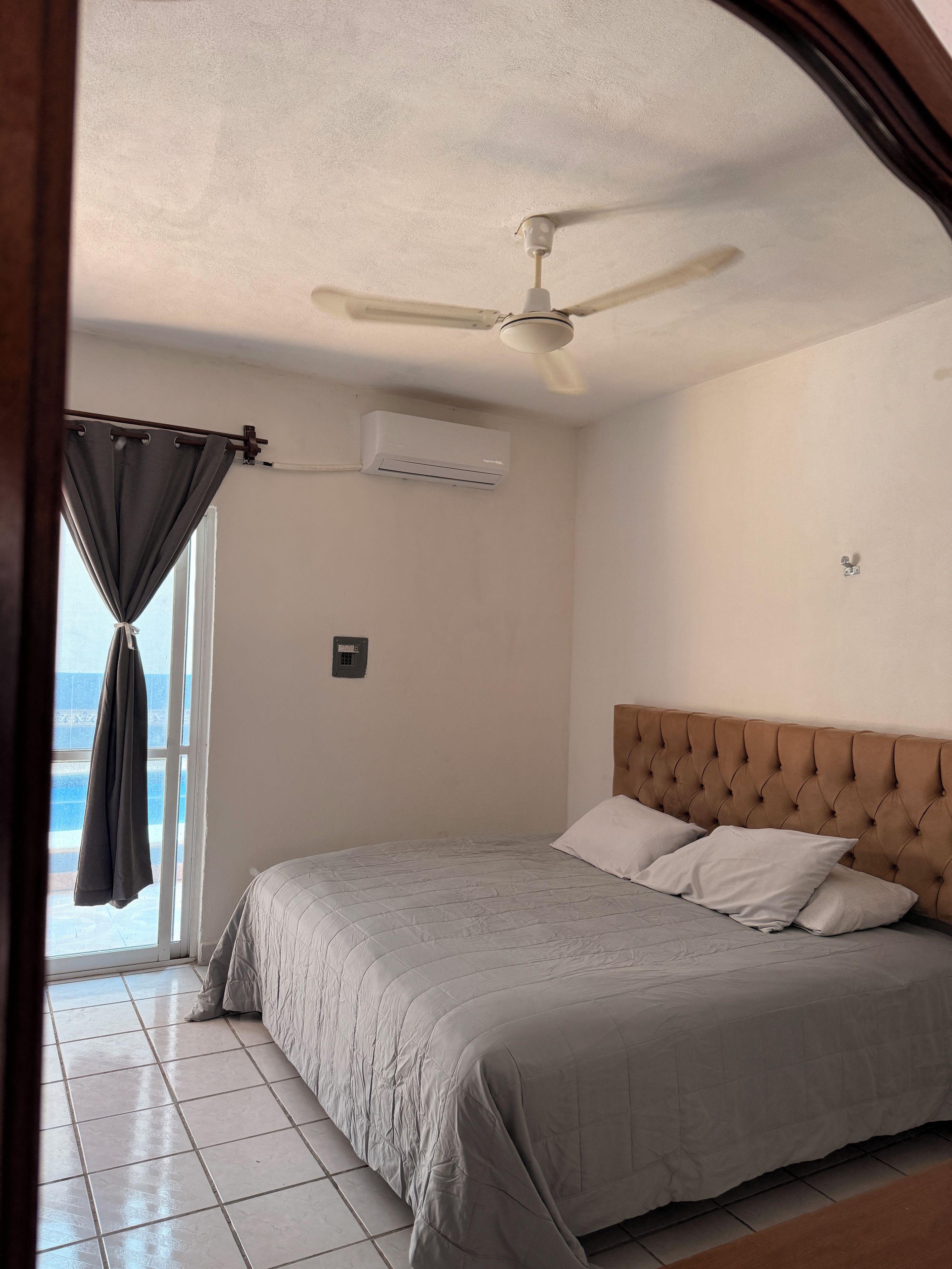 Une chambre à coucher comprenant un lit, un ventilateur de plafond, un appareil de climatisation, une fenêtre avec des rideaux et un plancher carrelé.