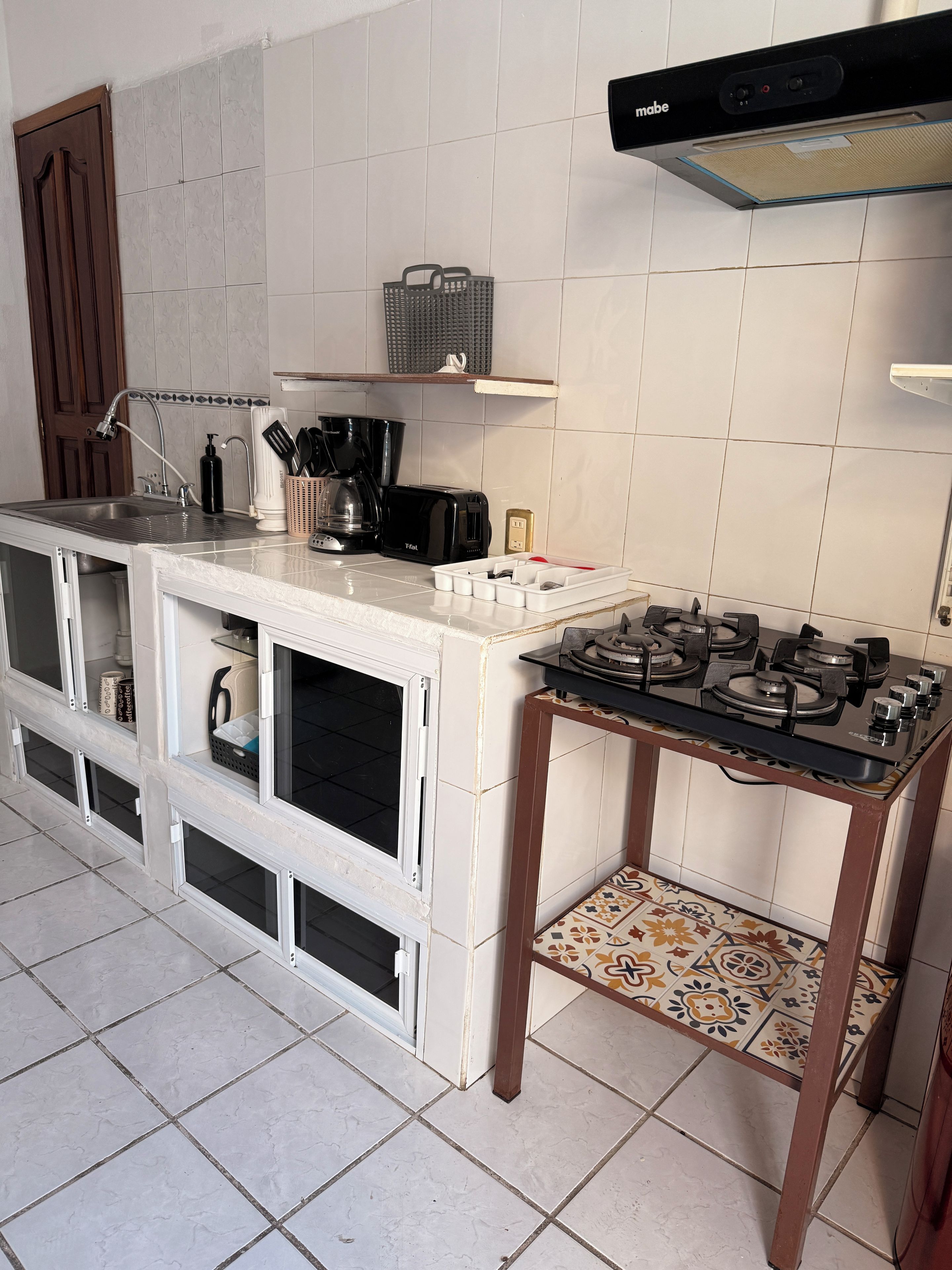 A kitchen with a gas stove, a sink, and a toaster. There is a shelf above the sink and a basket on the wall.