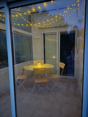 Een glazen patio met een ronde tafel en stoelen, sfeerverlichting en een bordje.