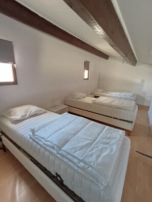 A room with three beds arranged in a row, wooden beams on the ceiling, and a small window.