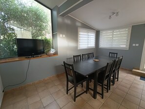 A dining area with a dark wooden table and chairs, a television on a window sill, and a view of greenery outside.