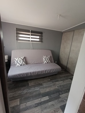 Ein Schlafzimmer mit grauem Sofa, gemusterten Kissen, einem Fenster mit Jalousien und einem Kleiderschrank.