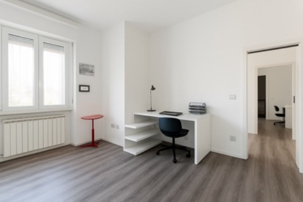 Ein Zimmer mit einem weißen Schreibtisch, einem schwarzen Bürostuhl und einem kleinen roten Tisch. Unter einem Fenster befindet sich eine Heizung, und eine Tür führt in einen anderen Raum.