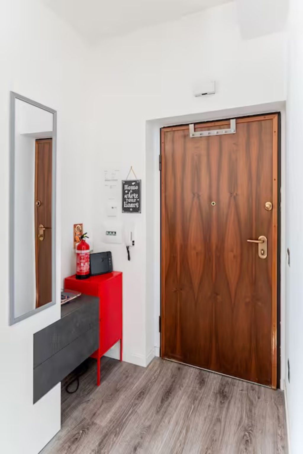 A hallway with a wooden door, a red cabinet, a fire extinguisher, and a wall-mounted light fixture.