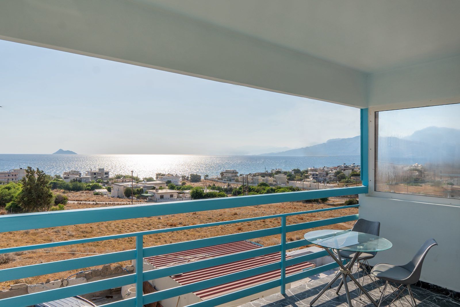 Un balcón con vistas a un pueblo costero, una mesa con una silla y un tapete a rayas.
