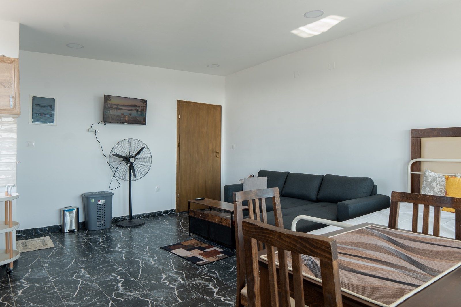 Een moderne woonkamer met een donkergrijze bank, houten stoelen, een eettafel, een ventilator en een kleine prullenbak.
