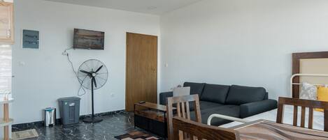 Een moderne woonkamer met een donkergrijze bank, houten stoelen, een eettafel, een ventilator en een kleine prullenbak.