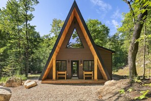A triangular wooden cabin with a front porch and two benches, surrounded by trees. - Spa & Nature | Celestra (Grenville-sur-la-Rouge)