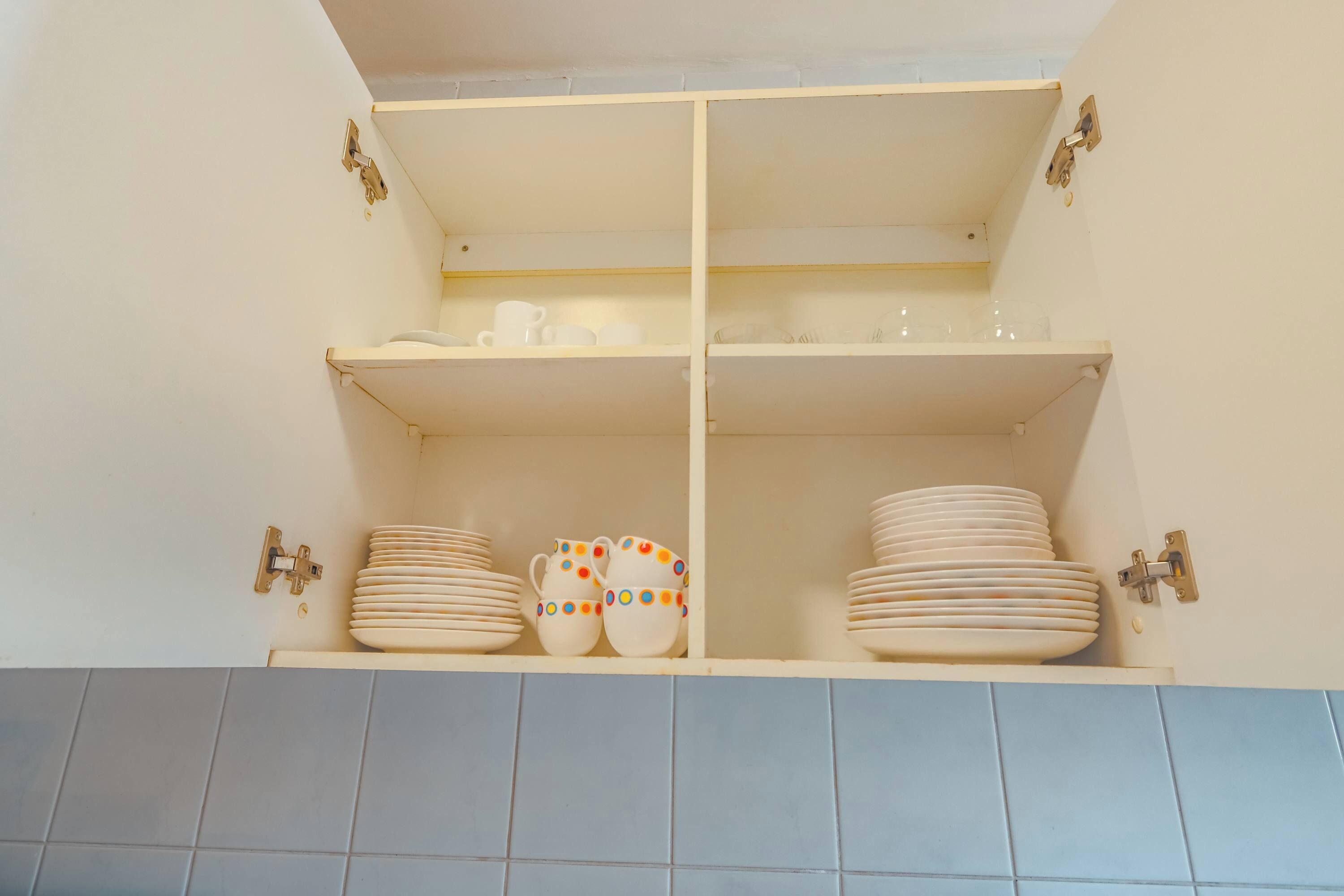 A kitchen cabinet with two shelves, each holding stacks of white plates and a pair of white cups with colorful polka dots.