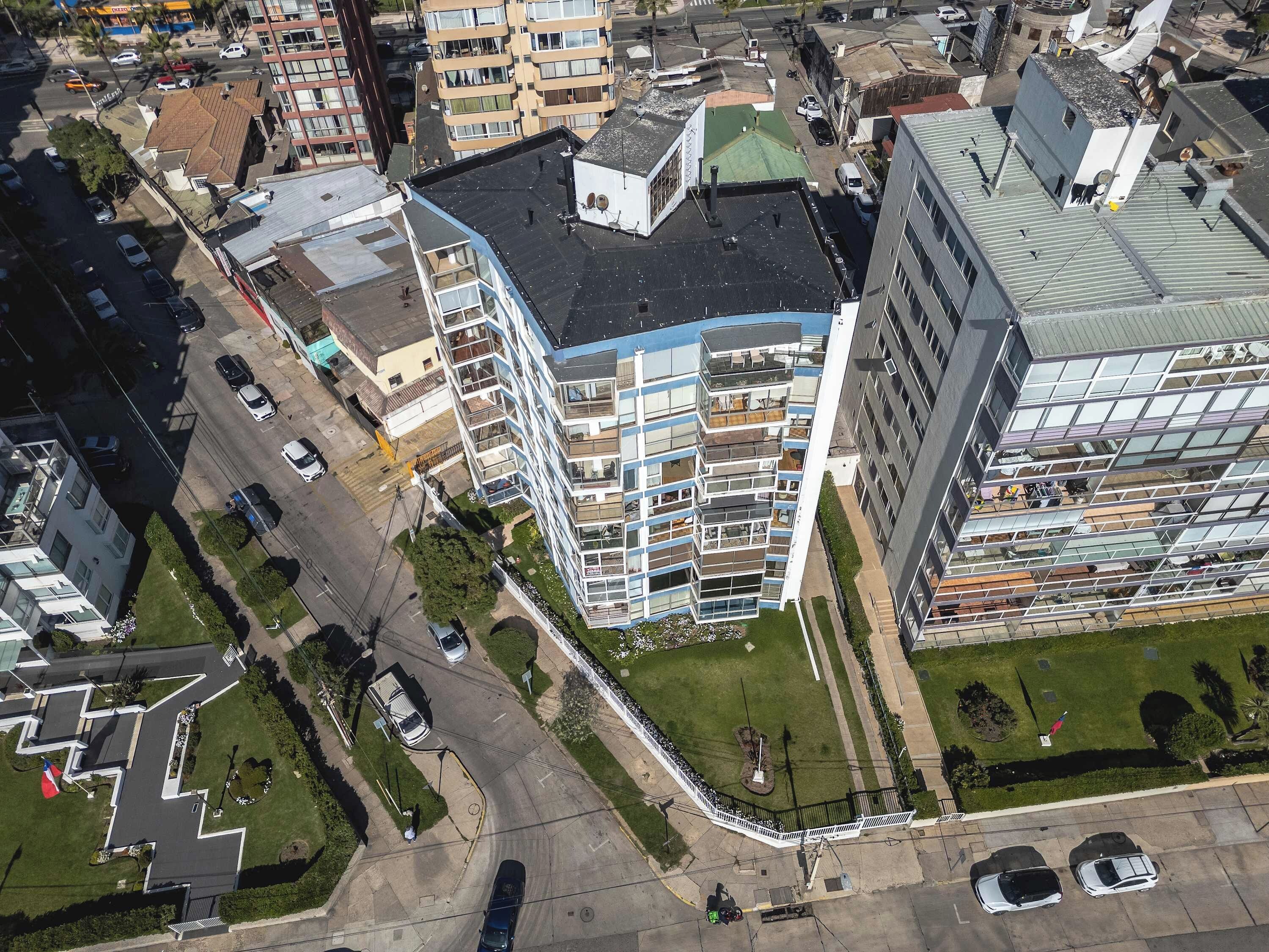 Un edificio de gran altura con varios balcones, zona de estacionamiento y un espacio verde con una bandera.